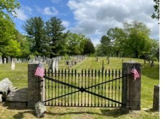 Iron cemetery gate with american flags on the right and left side, cemetery gravestones in rows with trees in the background
